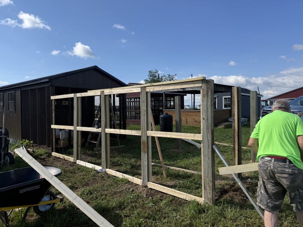 Man in a bright green shirt stands beside a wooden framing structure outdoors under a clear blue sky; farm buildings and fencing in the background.
